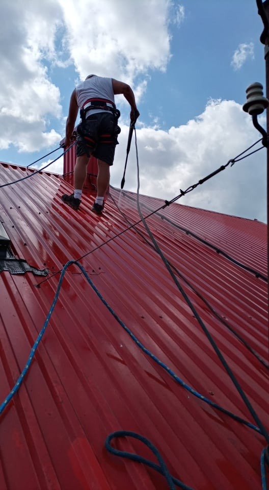 Pracownik w uprzęży, z perspektywy żaby, myje czerwoną blachodachówkę na dachu za pomocą lancy ciśnieniowej, zabezpieczony linami.