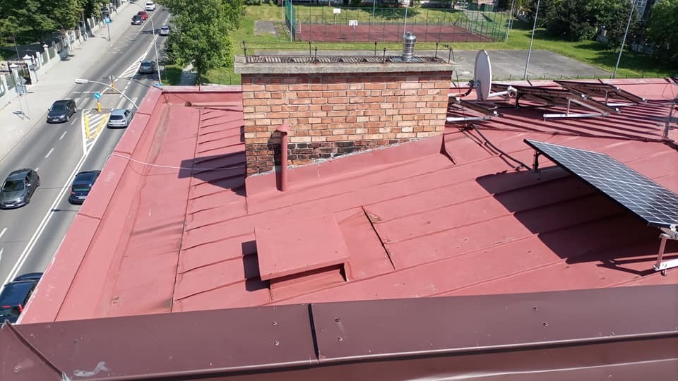 Czerwony blaszany dach z ceglanym kominem, panelami słonecznymi i widokiem na ulicę z samochodami w słoneczny dzień.