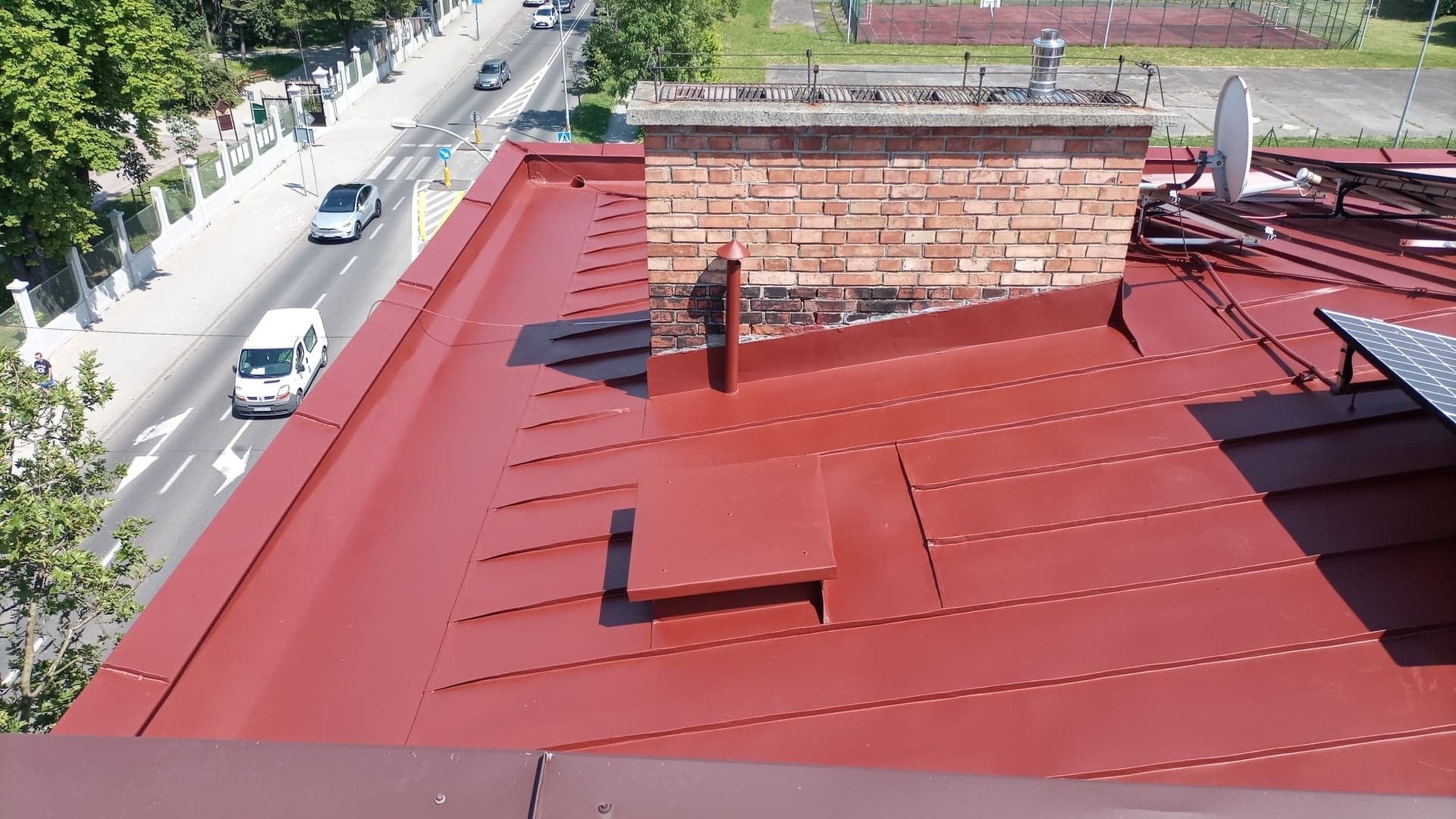 Czerwony, metalowy dach z kominem, anteną satelitarną i panelami słonecznymi widziany z góry, z widokiem na ulicę i budynki w tle.