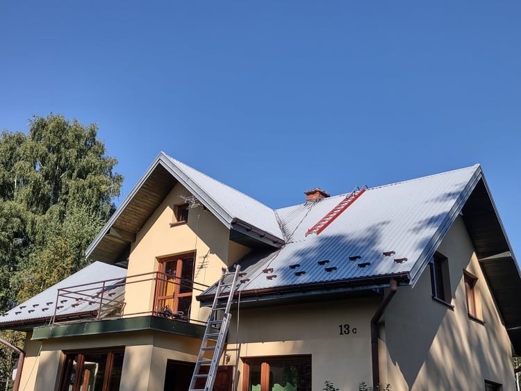 Dwupiętrowy dom z jasną elewacją i metalowym dachem, na którym znajduje się drabina dekarska z czerwonymi szczeblami, oparta o komin.