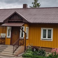 Żółty dom z brązowym dachem i rynnami, widok z zewnątrz, kwiaty w ogródku przed wejściem.