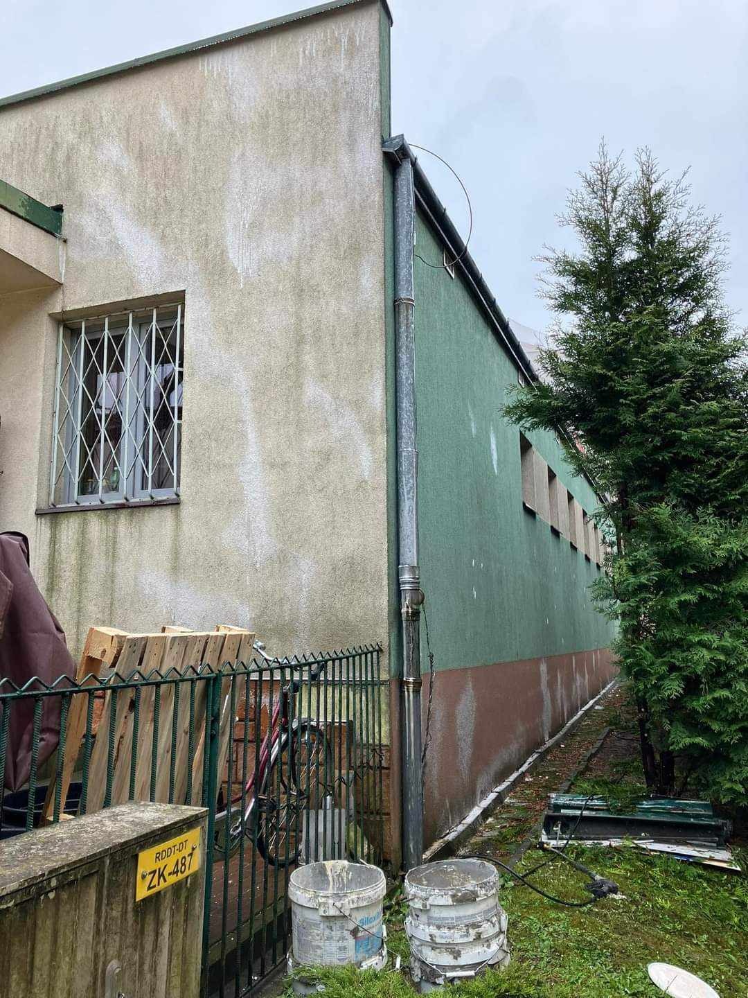 Dwukolorowa elewacja budynku z widocznymi zabrudzeniami i zaciekami, rynna, drewniane palety oparte o ogrodzenie, rower i dwa wiaderka po farbie na trawniku.