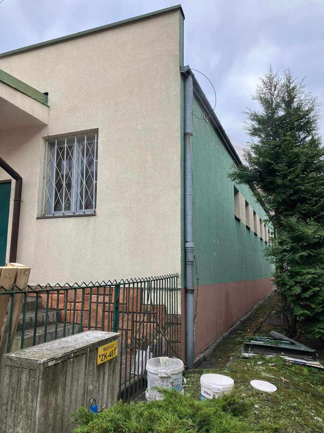 Dwukolorowa elewacja budynku w Luszowicach, z widocznymi wiadrami farby i schodami z cegły, częściowo zasłonięta przez zieleń.