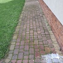 Zaniedbany chodnik z kostki brukowej, miejscami porośnięty mchem, przylegający do ściany budynku z ceglaną okładziną i trawnika.