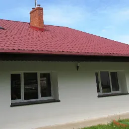 Boczny widok domu jednorodzinnego z czerwonym dachem, kominem z cegły, anteną satelitarną i dwoma oknami z białymi ramami na białej ścianie.