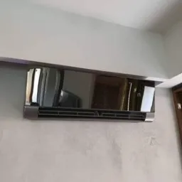 Błyszczący, czarny klimatyzator ścienny zamontowany w rogu pomieszczenia z widocznym fragmentem telewizora w odbiciu.