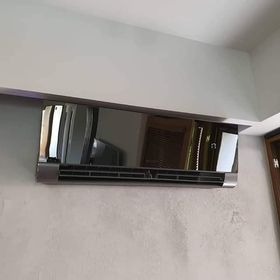 Błyszczący, czarny klimatyzator ścienny zamontowany w rogu pomieszczenia z widocznym fragmentem telewizora w odbiciu.