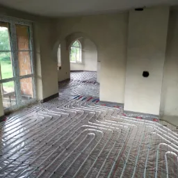Rozłożona instalacja ogrzewania podłogowego w surowym stanie, widoczne białe rurki na folii z czerwoną siatką, wnętrze z oknami i łukowymi przejściami.