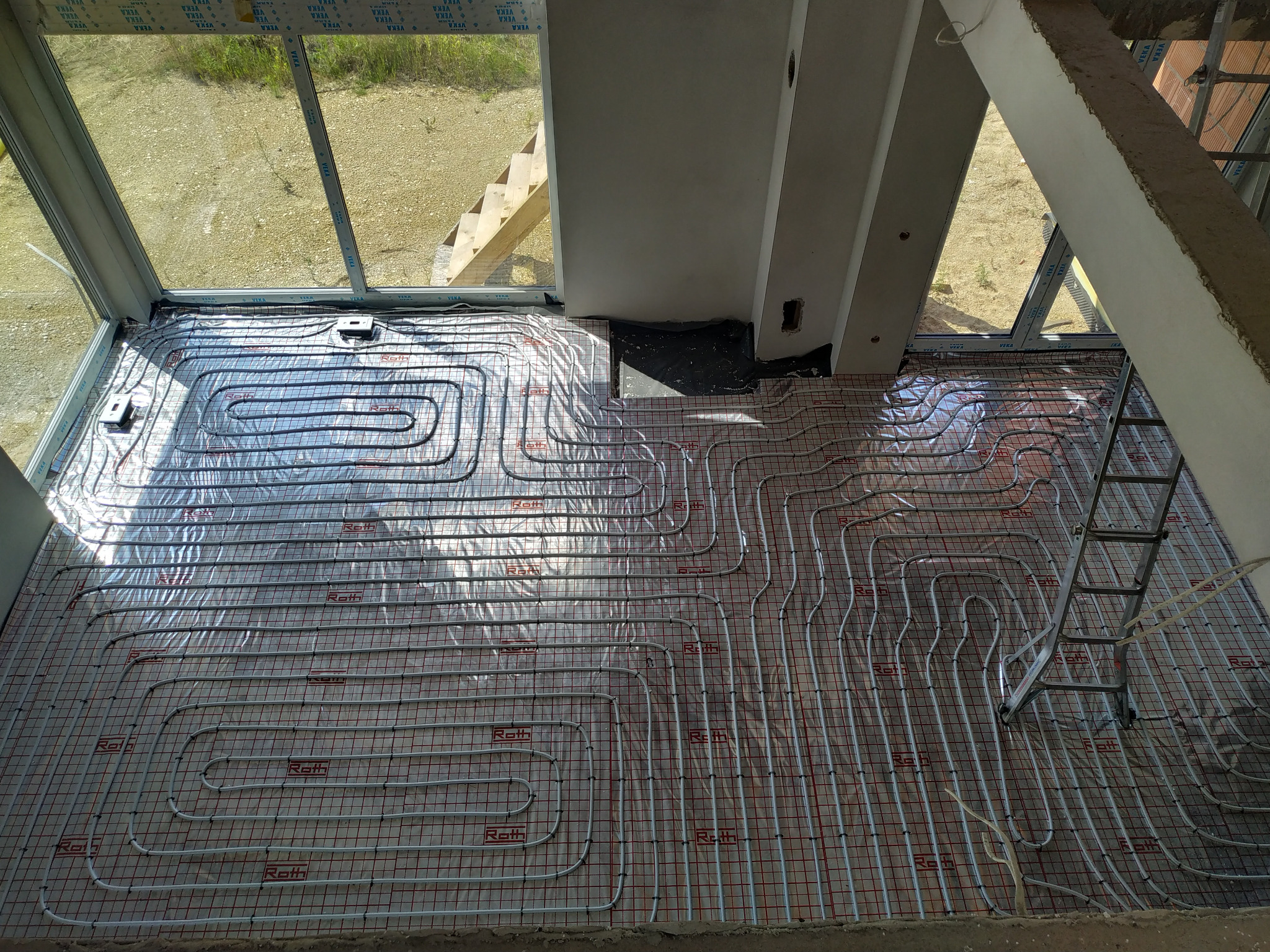 Rozłożone rury ogrzewania podłogowego marki Roth na izolacji z siatką w surowym wnętrzu domu w trakcie budowy, widok z góry na dużą powierzchnię, drabina oparta o ścianę, okno z widokiem na zewnątrz.