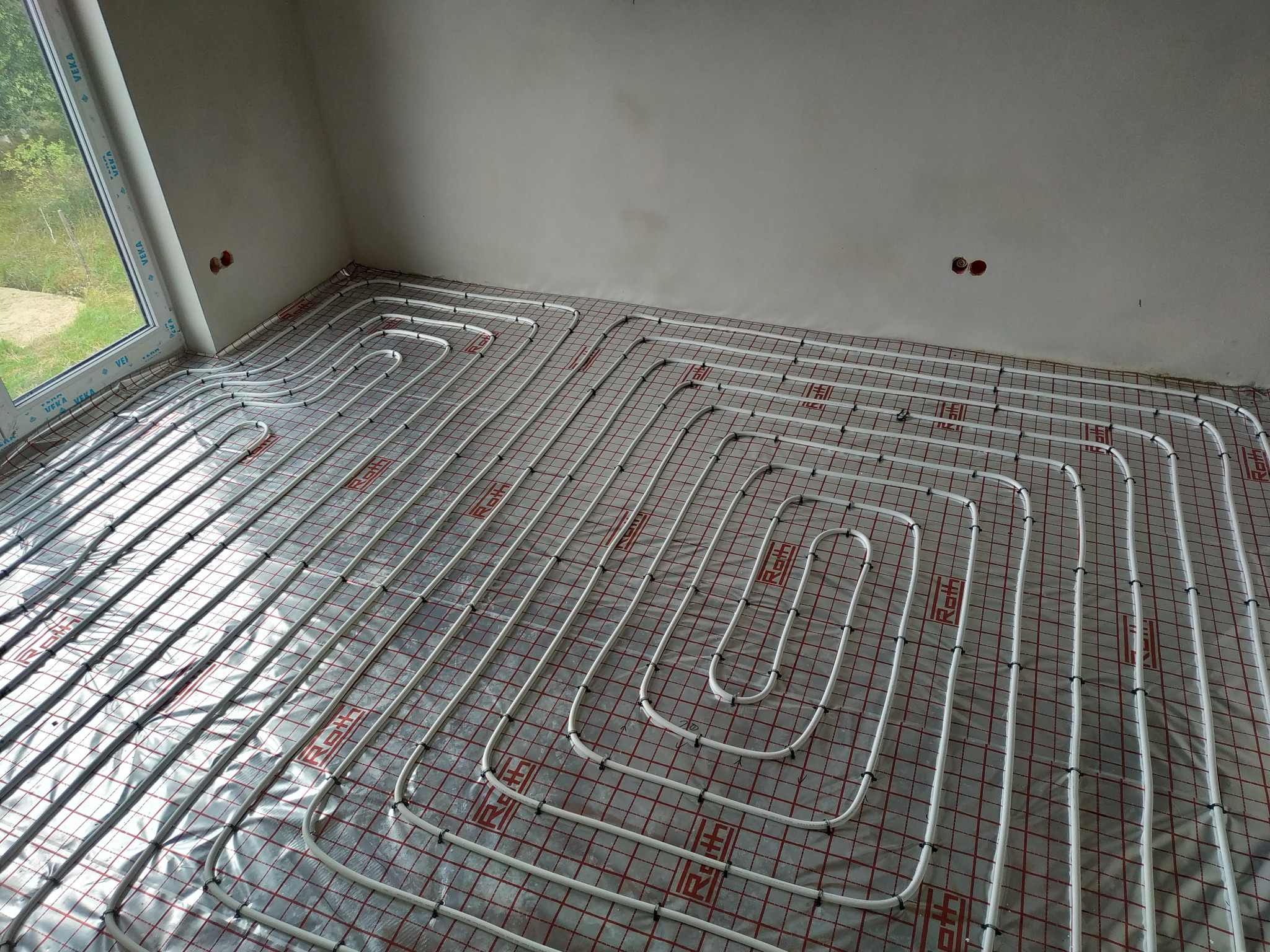 Białe rurki ogrzewania podłogowego ułożone w pętle na folii z czerwoną siatką, widoczne czarne zaciski mocujące, surowy stan wykończenia wnętrza z oknem w tle.