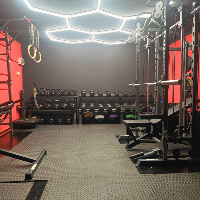 Profesjonalna siłownia domowa z czarnymi ścianami, czerwonymi akcentami i nowoczesnym oświetleniem LED. Widoczne kettlebells, maszyna Smitha i podłoga z gumy.
