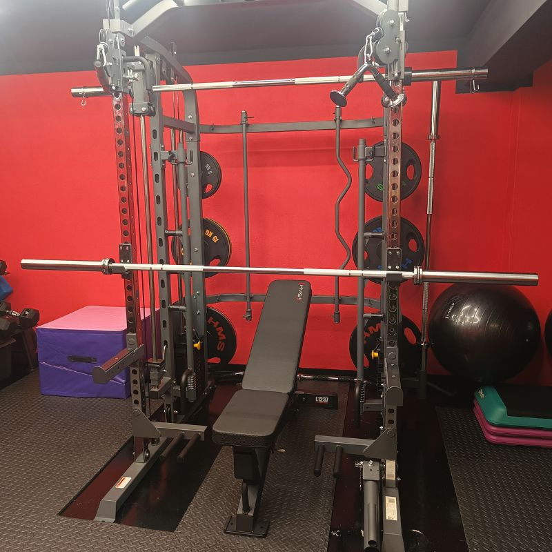 Profesjonalna klatka Smitha z obciążeniami, ławeczką i piłką do ćwiczeń na tle czerwonej ściany w siłowni. Sprzęt HMS z widocznymi detalami.