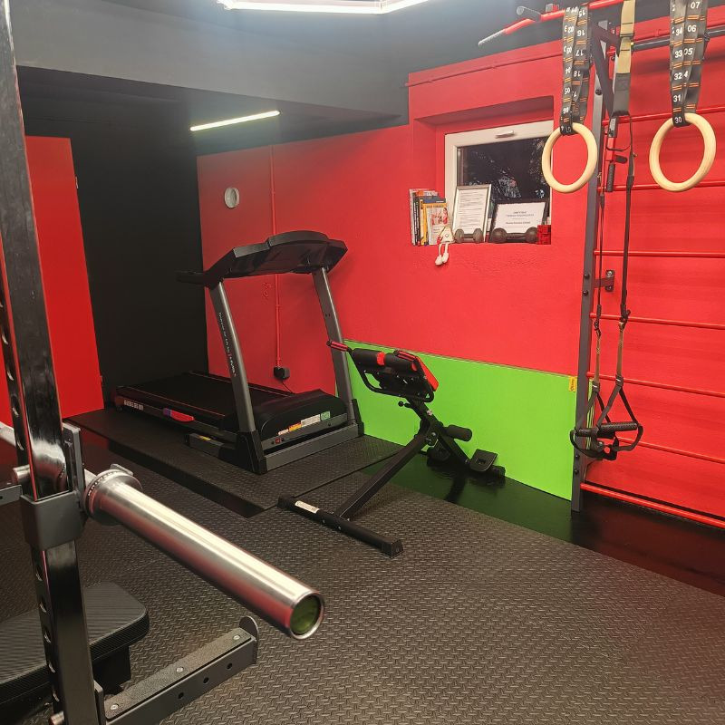 Profesjonalna siłownia domowa z bieżnią, rowerem stacjonarnym i drążkiem do podciągania na tle czerwonej ściany z oknem i zielonym akcentem.
