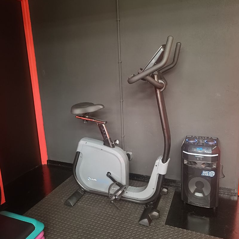Szary rower treningowy pionowy z czarnym siedziskiem i kierownicą, stojący na czarnej macie w siłowni, obok głośnik Bluetooth. Sprzęt do ćwiczeń w pomieszczeniu.