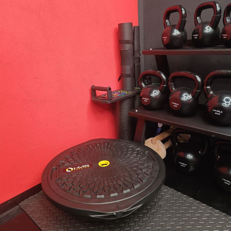 Czarny balansujący dysk HMS na podłodze siłowni, obok półki z kettlebells i czerwonej ściany. Sprzęt do ćwiczeń równowagi i stabilizacji, widok z boku.