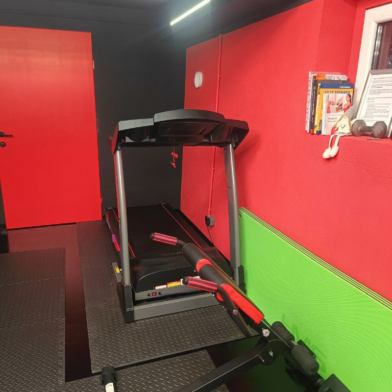 Czerwono-czarna siłownia domowa z bieżnią, książkami o treningu i hantlami na półce. Ściany w intensywnych kolorach, podłoga z czarnej gumy.