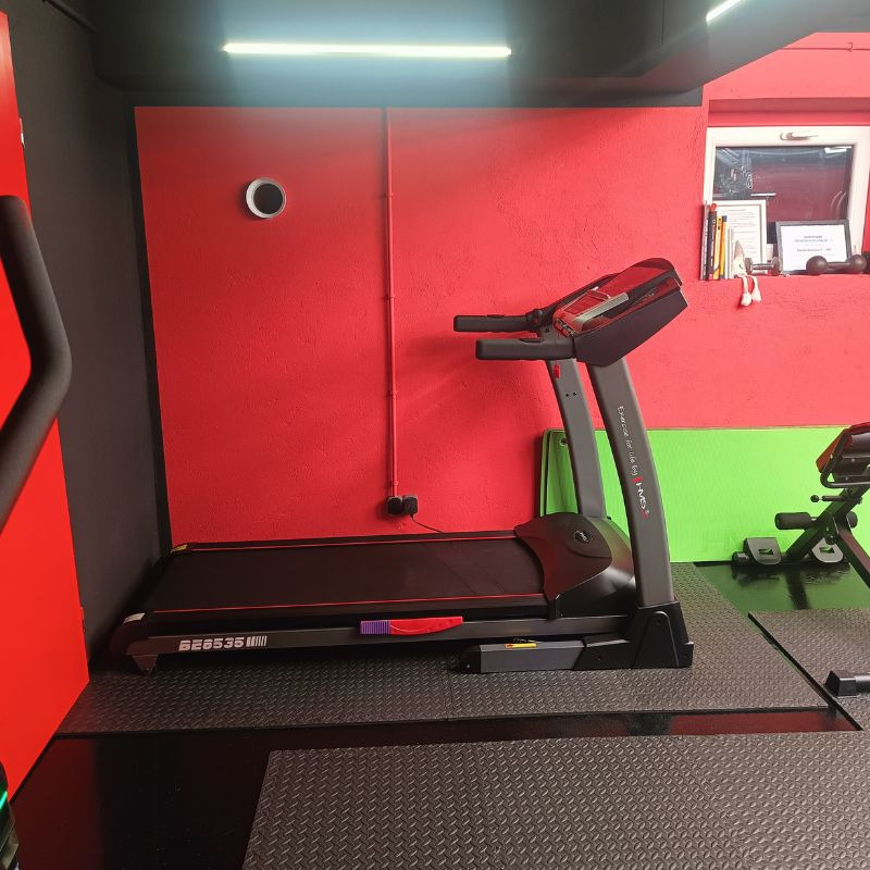 Bieżnia w siłowni w Miłomłynie, na tle czerwonej ściany z oknem. Widoczny sprzęt do ćwiczeń i podłoga z gumowej kostki. Oświetlenie sufitowe.