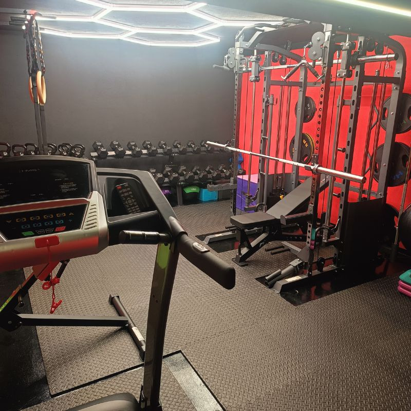 Profesjonalna siłownia domowa z bieżnią, sztangą, hantlami i drabinkami gimnastycznymi. Ciemne ściany, jasne oświetlenie sufitowe.