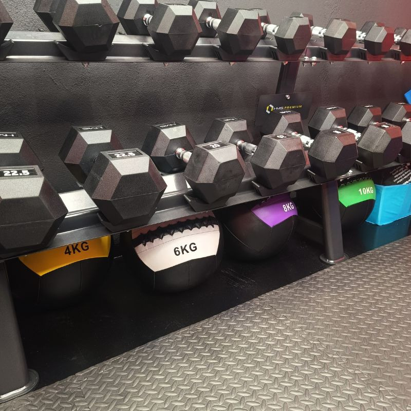 Regał z hantlami i piłkami lekarskimi o różnych wagach w siłowni. Hantle w górnych rzędach, piłki lekarskie w dolnym rzędzie. Podłoga z antypoślizgową fakturą.