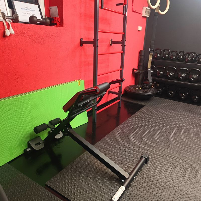 Wyposażenie siłowni: ławka rzymska, drabinka gimnastyczna, kettlebells. Czerwona ściana i czarna podłoga z gumy. Sprzęt do ćwiczeń w nowoczesnym wnętrzu.