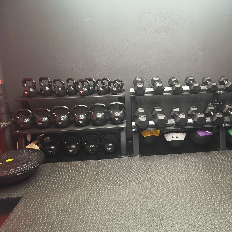 Uporządkowany zestaw czarnych kettlebell i hantli na metalowych regałach w siłowni, z widoczną podłogą z gumy i piłkami lekarskimi w dolnej części kadru.