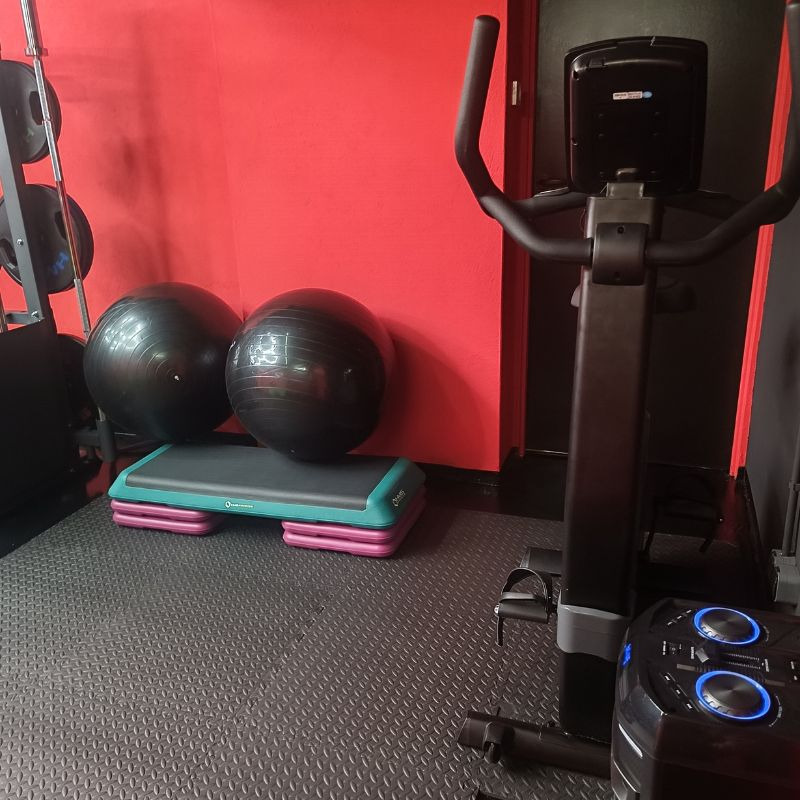 Sprzęt do ćwiczeń: piłki, step i rower stacjonarny na tle czerwonej ściany w siłowni. Widoczny fragment podłogi z gumowej maty.