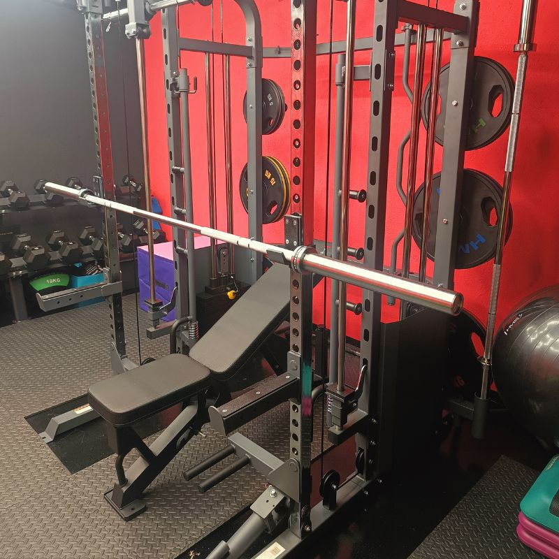 Profesjonalny sprzęt do ćwiczeń siłowych: suwnica Smitha z ławką, obciążeniami i hantlami w tle, na tle czerwonej ściany. Nowoczesna siłownia.