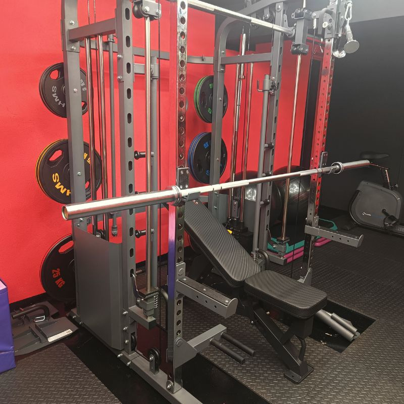Profesjonalna klatka Smitha z obciążeniami i ławką w siłowni, czerwona ściana z dyskami, czarna podłoga, sprzęt do ćwiczeń siłowych.