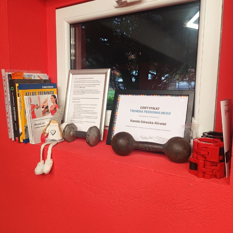 Certyfikat trenera personalnego Kamili Góreckiej-Kirwiel, książki o treningu, hantle i akcesoria na czerwonym parapecie okna. W tle widok z okna.