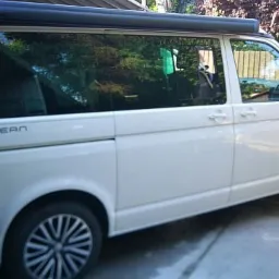 Biały samochód typu van z zamontowaną markizą przeciwsłoneczną, model Ocean, zaparkowany na betonowym podjeździe przed ogrodzeniem.