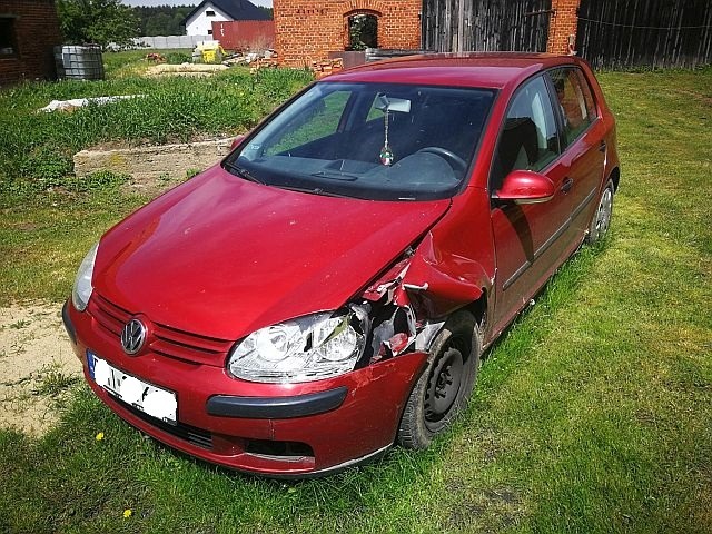 Czerwony Volkswagen Golf z uszkodzonym przodem stoi na trawie, widoczne zgniecione błotniki i uszkodzony reflektor.