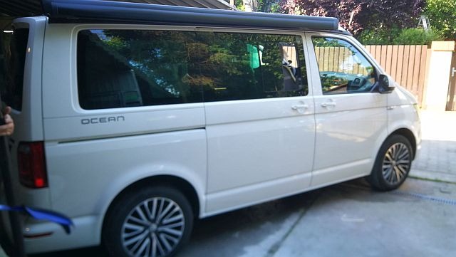 Biały samochód typu van z zamontowaną markizą przeciwsłoneczną, model Ocean, zaparkowany na betonowym podjeździe przed ogrodzeniem.