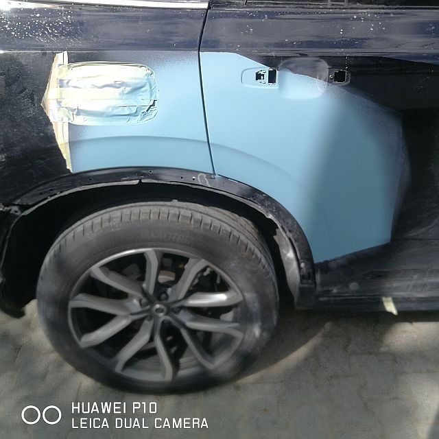 Fragment samochodu w trakcie naprawy blacharsko-lakierniczej: boczny panel i wnęka w kolorze jasnoniebieskim, zabezpieczone taśmą, widoczne koło i czarny fragment karoserii.