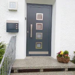 Szara, nowoczesna brama wejściowa z czterema okienkami i długim, pionowym uchwytem, zamontowana w białej, otynkowanej ścianie. Przed wejściem granitowe schody z koszem bratków i skrzynka na listy.