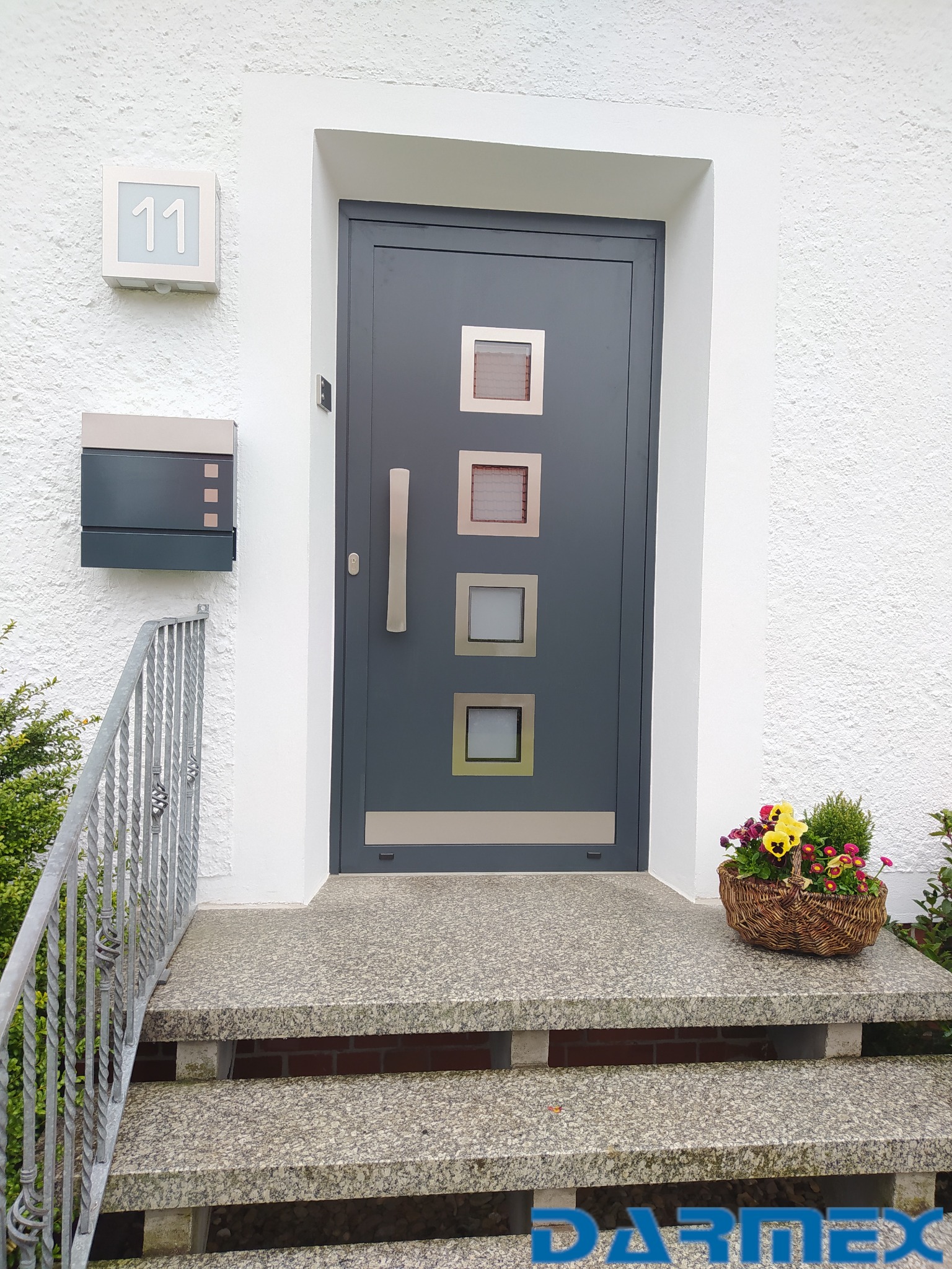 Szara, nowoczesna brama wejściowa z czterema okienkami i długim, pionowym uchwytem, zamontowana w białej, otynkowanej ścianie. Przed wejściem granitowe schody z koszem bratków i skrzynka na listy.