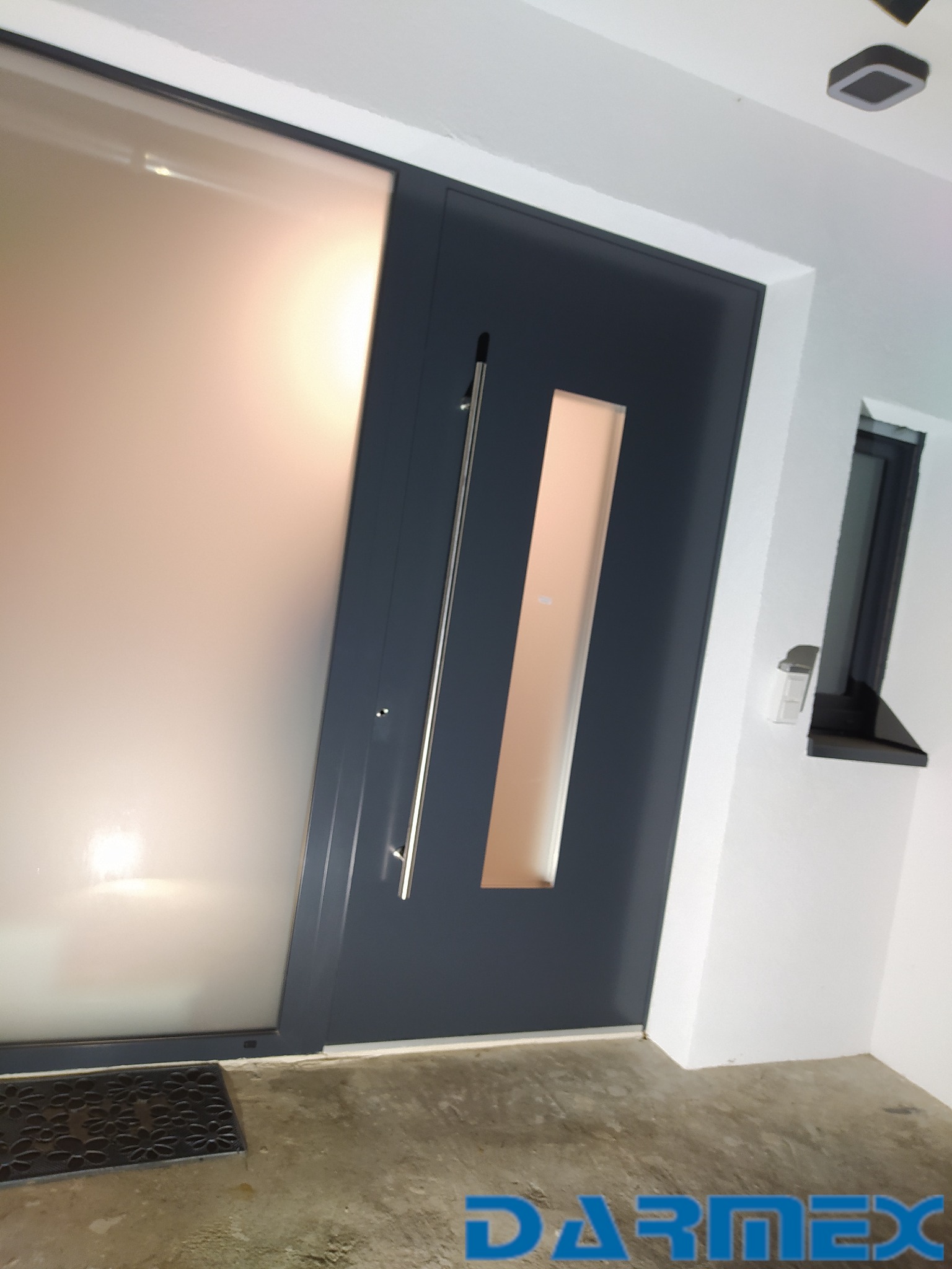 Nowoczesne drzwi wejściowe z panelem z matowego szkła i długim, pionowym uchwytem, z widocznym fragmentem progu i wycieraczki. W tle widoczne okno.