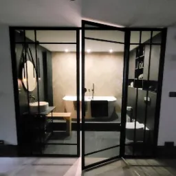 Nowoczesna łazienka z wanną wolnostojącą, umywalką nablatową i kabiną prysznicową z czarnymi, metalowymi profilami, widziana przez otwarte drzwi z podobnymi profilami.