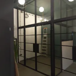 Wnętrze z przeszkloną ścianą działową z czarnymi metalowymi profilami, widoczna drabina aluminiowa i elementy oświetlenia.