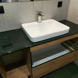 Łazienka z umywalką nablatową o kwadratowym kształcie, chromowaną baterią i czarnym blatem z widoczną strukturą kamienia, szafka pod umywalką z drewnopodobnym wykończeniem i otwartą półką.