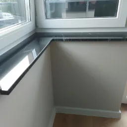 Czarny parapet z konglomeratu zamontowany w narożnym oknie, widok z bliska, jasne ściany i drewnopodobna podłoga.