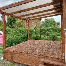 Drewniany taras z zadaszeniem z poliwęglanu, oświetlony sznurem żarówek, z widokiem na zarośnięty ogród i ceglany budynek w tle, z detalami na stole: telefon, kubek i akcesoria.