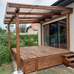 Zadaszony drewniany taras z podestem i schodami, oświetlony girlandą żarówek, z widokiem na ogród i dom w tle.