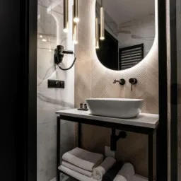 Nowoczesna łazienka z owalnym lustrem podświetlanym LED, wiszącymi lampami w kształcie złotych tub, białą umywalką nablatową i marmurowymi płytkami na ścianach.