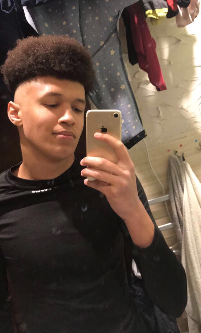 Młody mężczyzna robi selfie telefonem, ubrany w czarną bluzkę, z wyraźnym cieniowaniem na twarzy i dekoracyjną ścianą w tle.