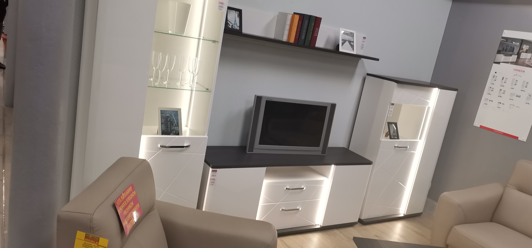 Nowoczesny zestaw mebli do salonu w odcieniach bieli i szarości, z oświetleniem LED, składający się z witryny, szafki RTV i półki wiszącej, uzupełniony elementami dekoracyjnymi i fotelami.