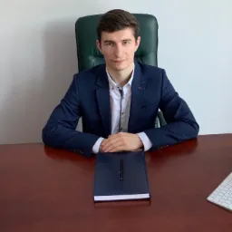 Młody mężczyzna w garniturze siedzi za biurkiem z notatnikiem, długopisem, klawiaturą i stosem książek prawniczych.