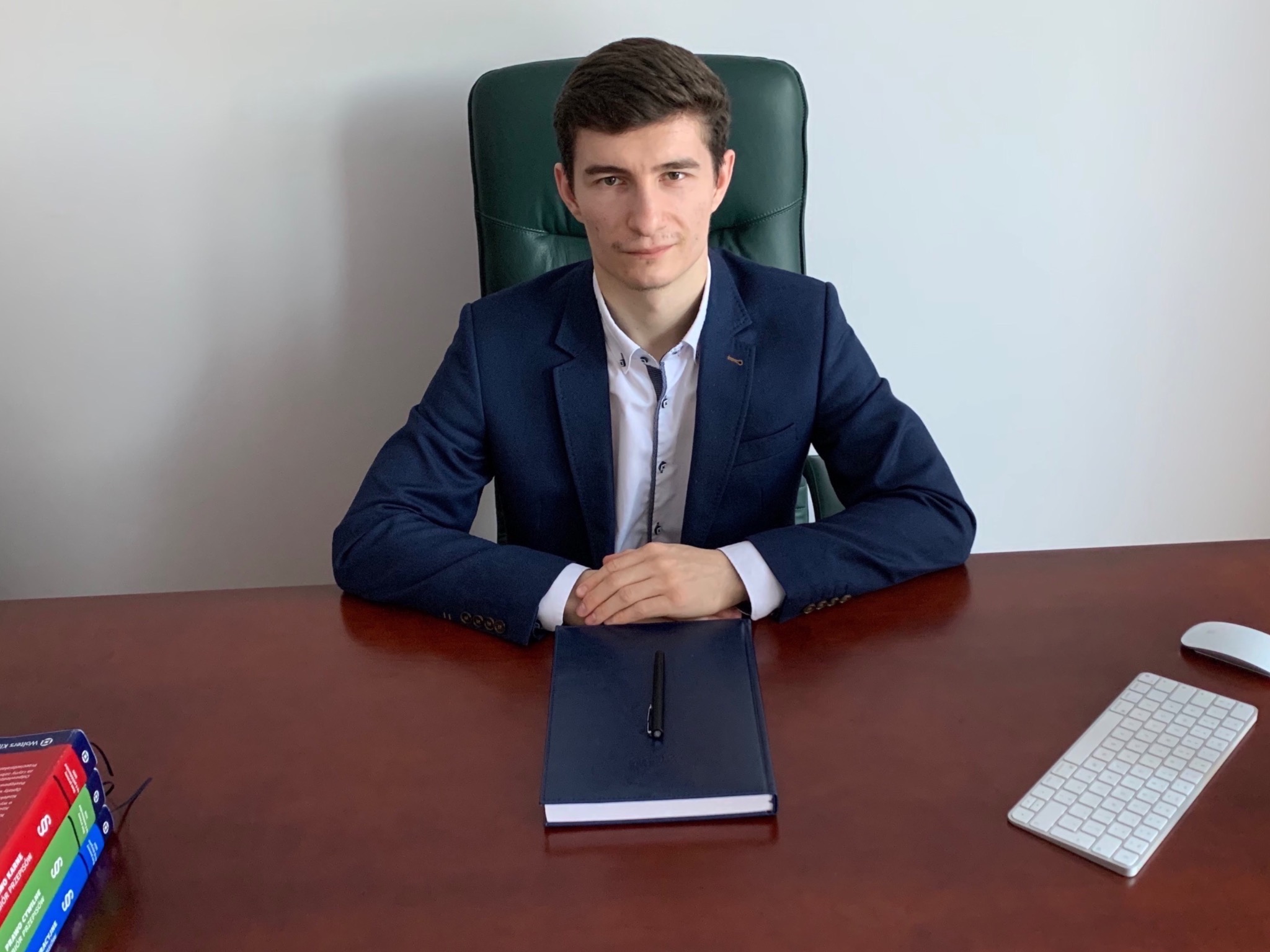 Młody mężczyzna w garniturze siedzi za biurkiem z notatnikiem, długopisem, klawiaturą i stosem książek prawniczych.