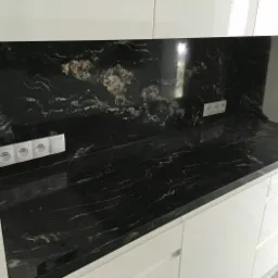 Czarny blat z kamienia o unikalnym wzorze z białymi żyłkami i złotymi plamami, dopasowany do ściany nad blatem, z widocznymi gniazdkami elektrycznymi i białymi szafkami kuchennymi.