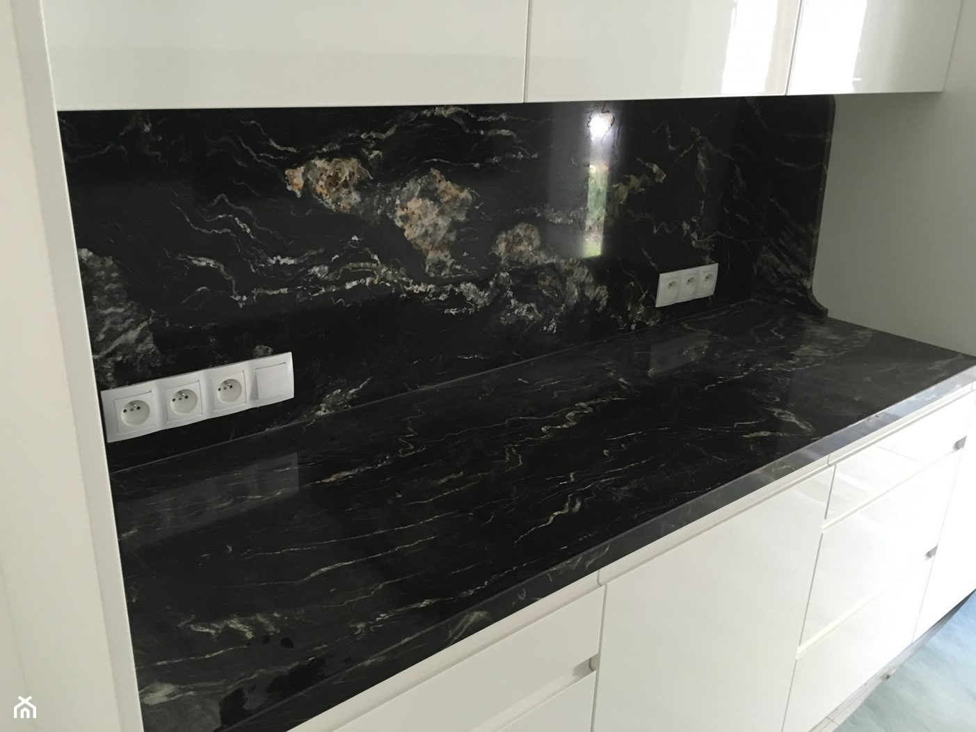 Czarny blat z kamienia o unikalnym wzorze z białymi żyłkami i złotymi plamami, dopasowany do ściany nad blatem, z widocznymi gniazdkami elektrycznymi i białymi szafkami kuchennymi.