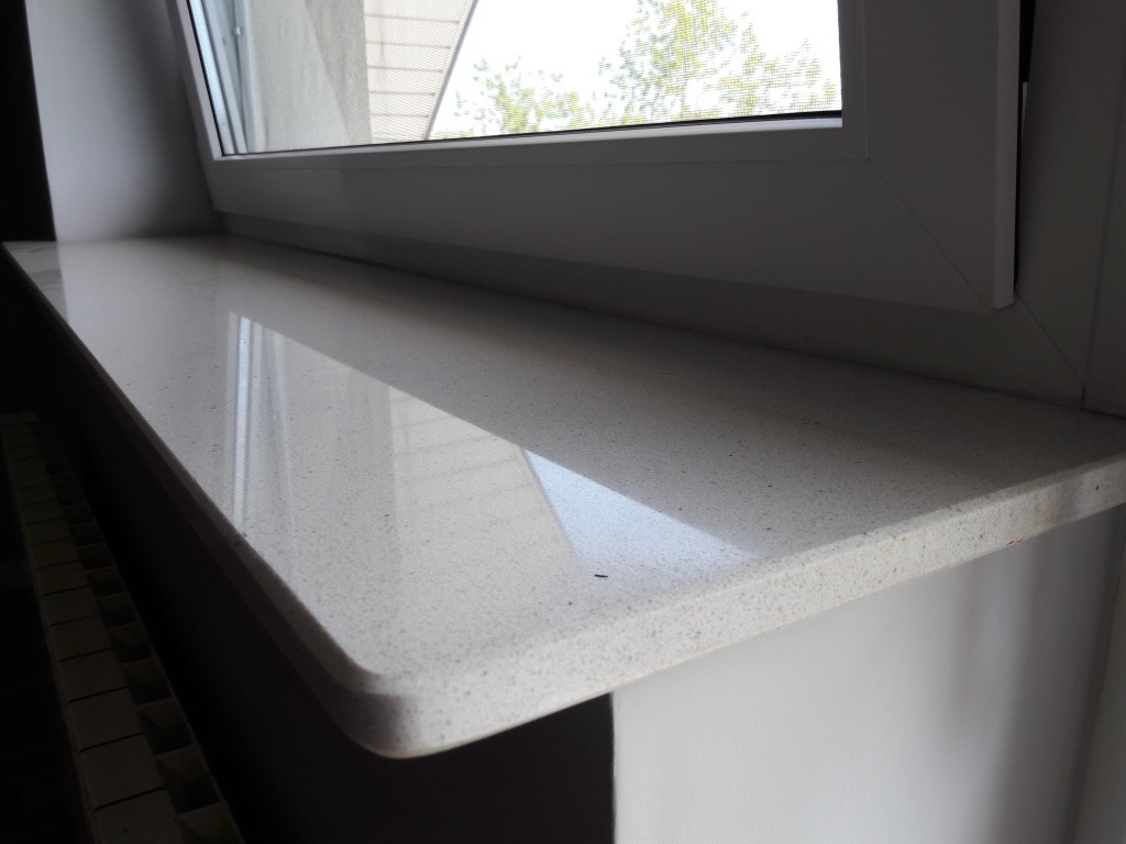 Jasny parapet z konglomeratu z zaokrąglonym brzegiem, zamontowany pod białym oknem, z widocznym fragmentem grzejnika.